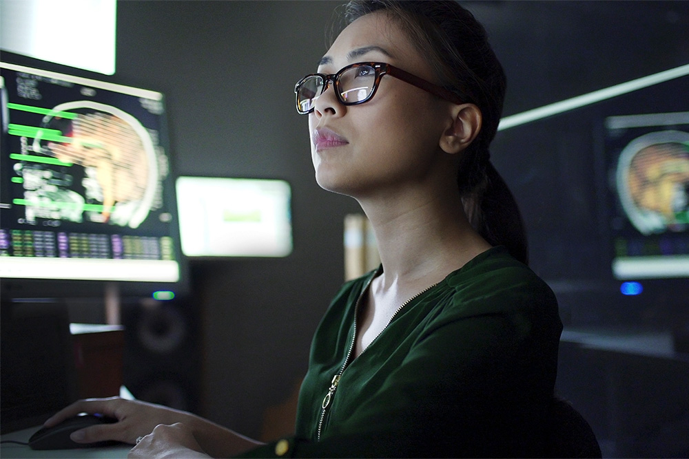 Close up stock image of a young asian woman working at a desk with multiple monitors. She’s studying the moving data & information on the screens which consists of human body scans, MRI & CAT, and scrolling text & numbers.