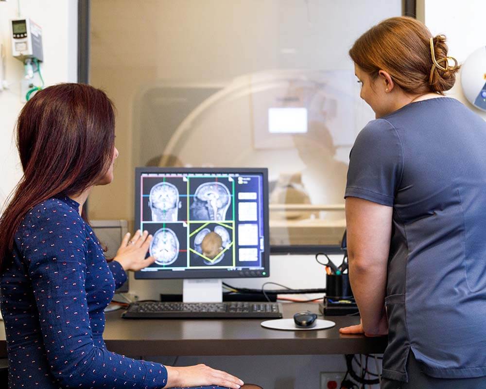 A doctor and a tech go over the results of an fMRI scan. You can see various images of a brain on the computer monitor in front of them and the fMRI machine in the room beyond the glass. The doctor sits and wears a long sleeved blue dress, she has dark straight hair. The tech stands and wears blue scrubs, she has light hair tied up on her head.